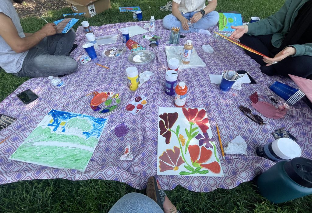 A purple blanket is laid out on green grass. There are paints, brushes, and small canvasses with partially finished paintings laid out on top. Along the edges of the blanket people can be seen from the shoulder down sitting cross legged . Two of them are holding small canvas boards and brushes in their hands.