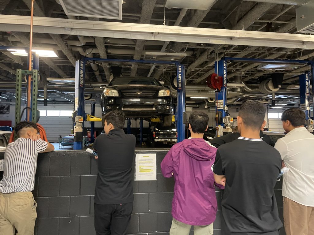 Six teenage boys are standing in a mechanic shop facing away from the camera. They are looking at a small black car that is set up on an auto lift. 