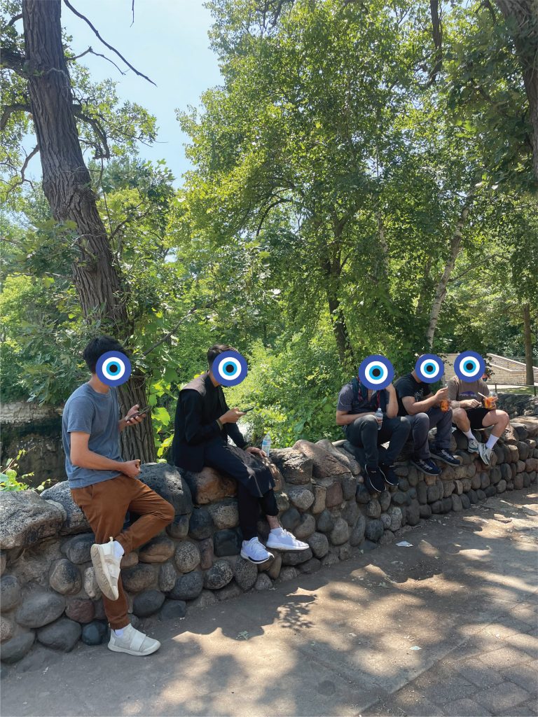 5 teenaged boys sit on a wall made of river stones. Behind the wall are tall trees, the sky is a clear blue.