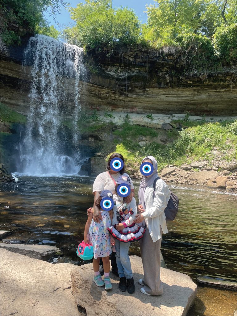 2 women and two young girls stand in front of a small waterfall on a sunny day.