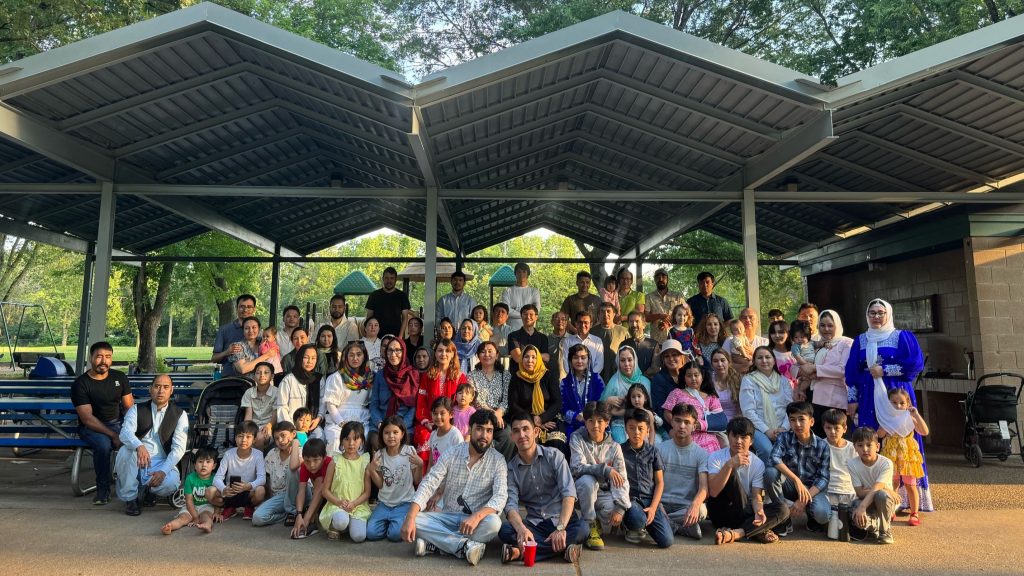 A large group of Hazara community members pose for a photo under an outdoor picnic area at a park.