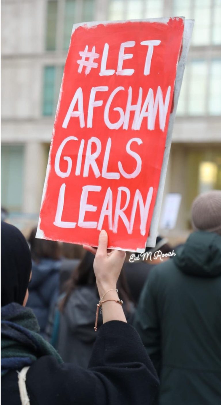 A red hand painted sign with white text is held above a crowd. It reads #LET AFGHAN GIRLS LEARN