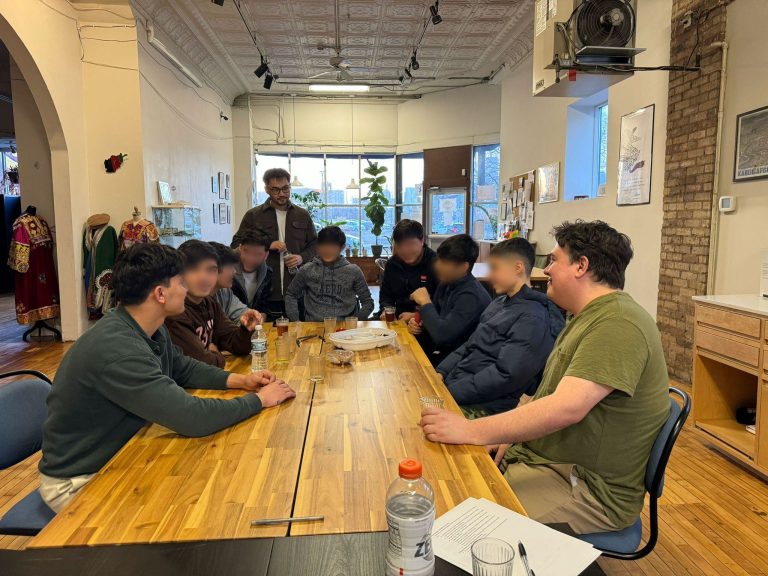 A group of boys sits around a wooden table with 3 adults, engaged in discussion.